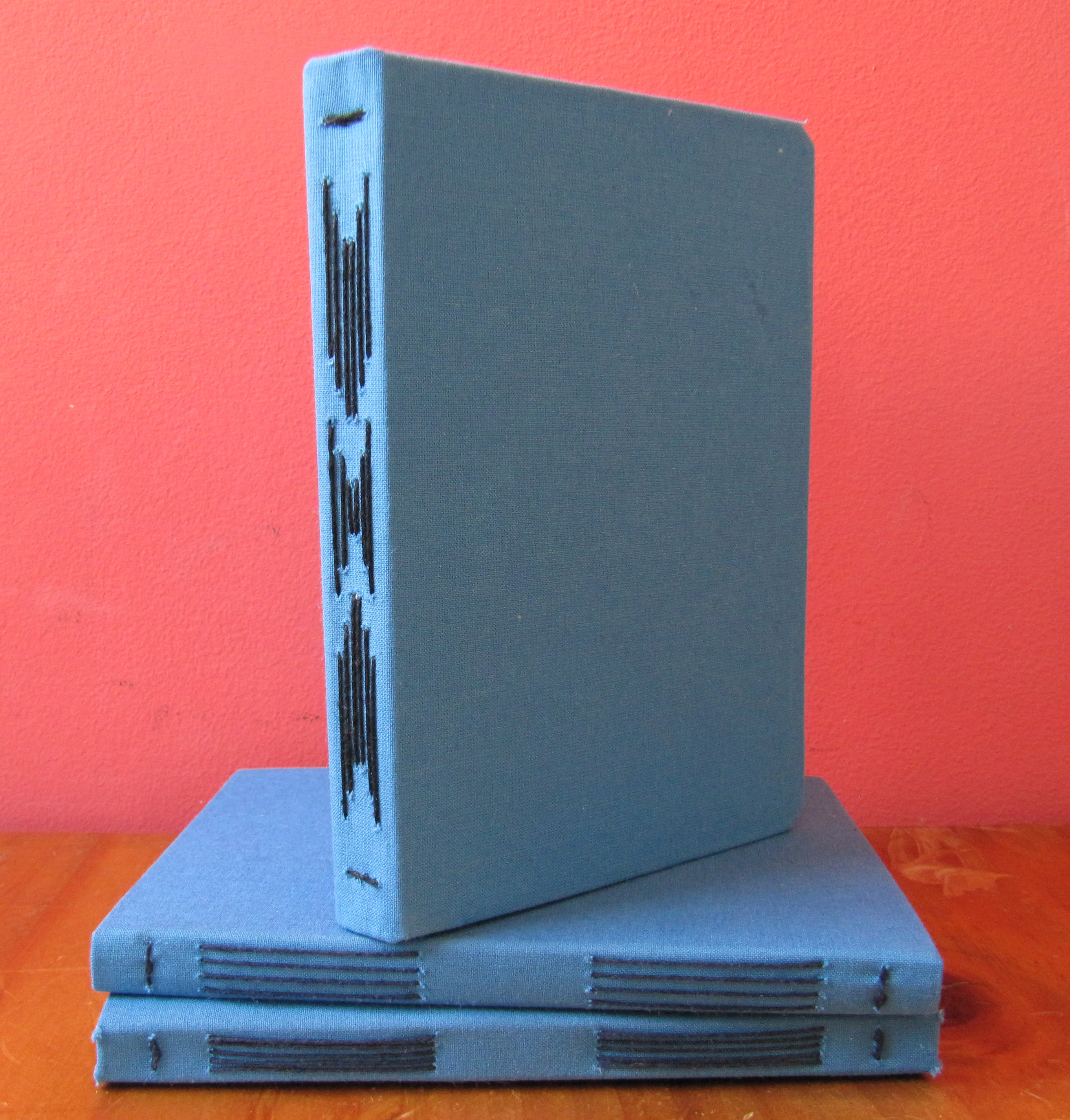 Three handmade sketchbooks stacked on top of each other; they have blue covers and are long stitch bound in multiple patterns in black thread.